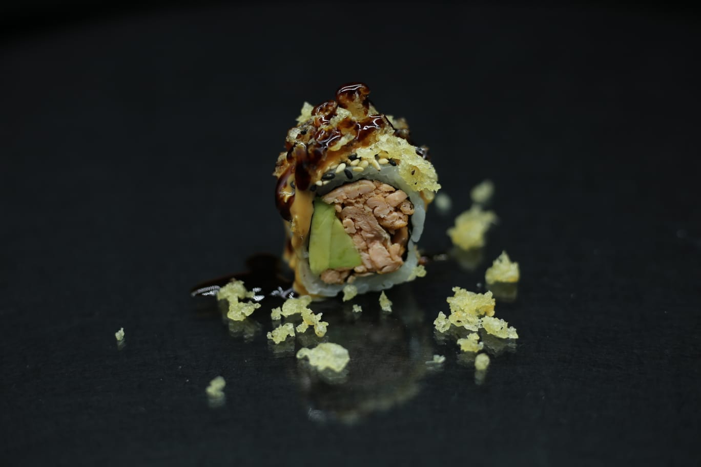 Primo piano di un rotolo di sushi su una superficie nera, ricoperto di salsa e circondato da briciole di riso sparse.
