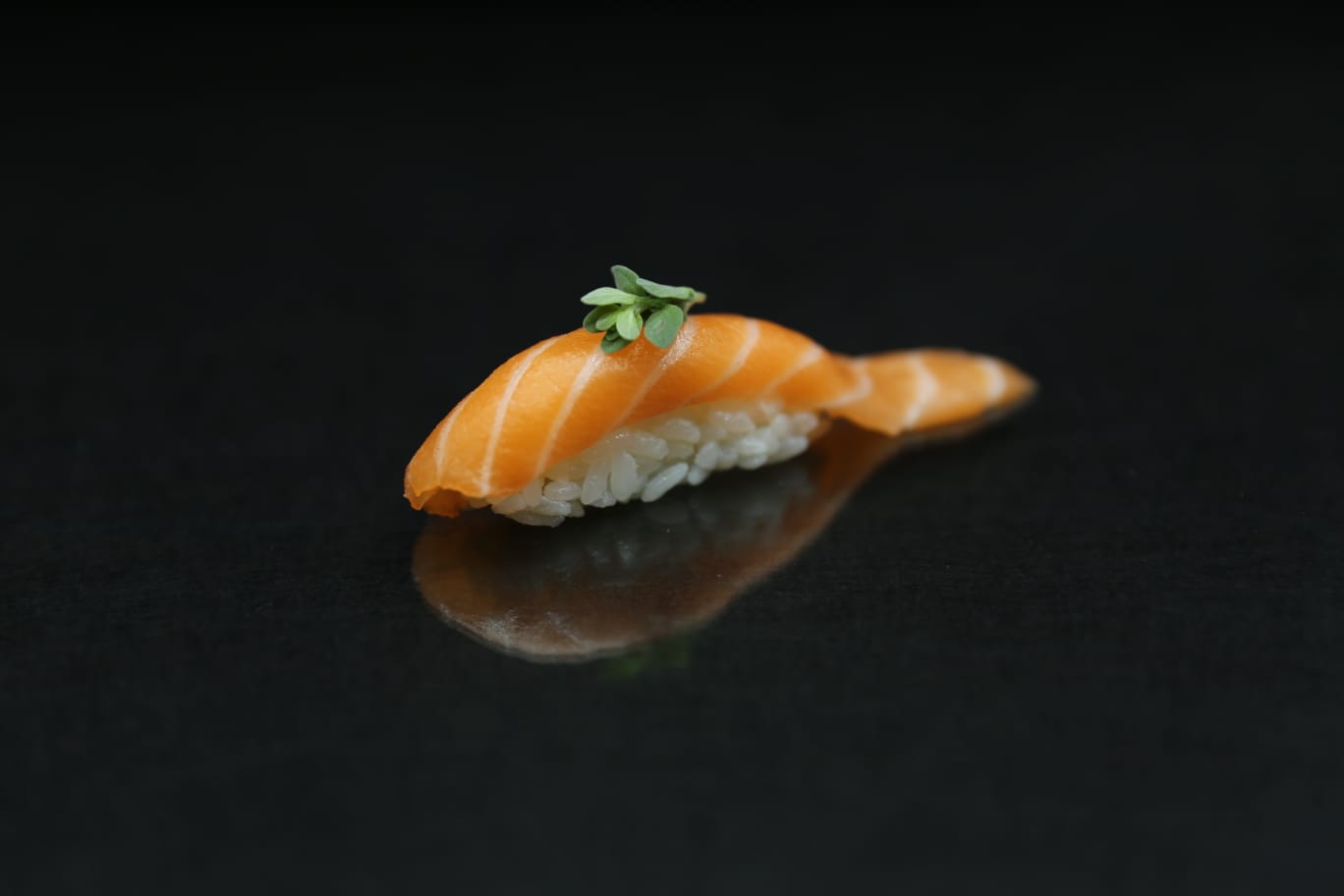 Sushi nigiri di salmone con guarnizione verde su una superficie scura e riflettente.