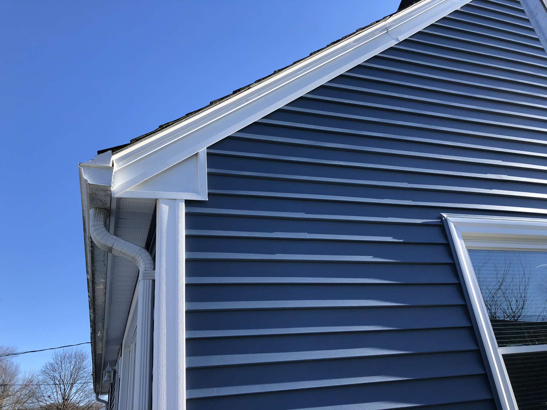A low-angle view of the corner of a house featuring blue horizontal siding, white trim, and a white downspout against blue sky.