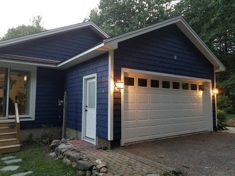 A blue house with a white garage door, a side entry door, and exterior lights, surrounded by trees and a stone path.