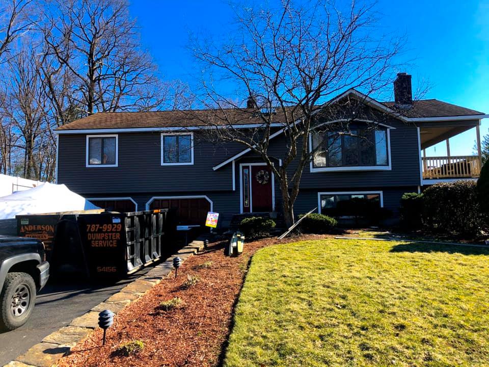 A gray split-level house with a red front door, a front lawn, and a large construction dumpster parked in the driveway.