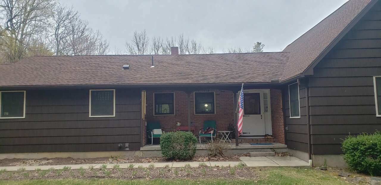 A one-story house with dark wood siding, a shingled roof, a front porch, and an American flag near the entryway.