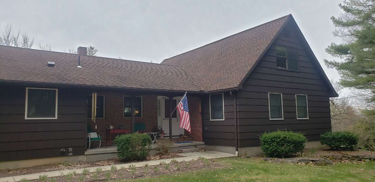A brown, single-story house with a steeply pitched roof, wood-style siding, and an American flag near the entryway.