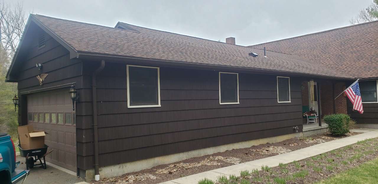 A side view of a dark brown wooden house with a single-car garage, three windows, and an American flag near the entrance.