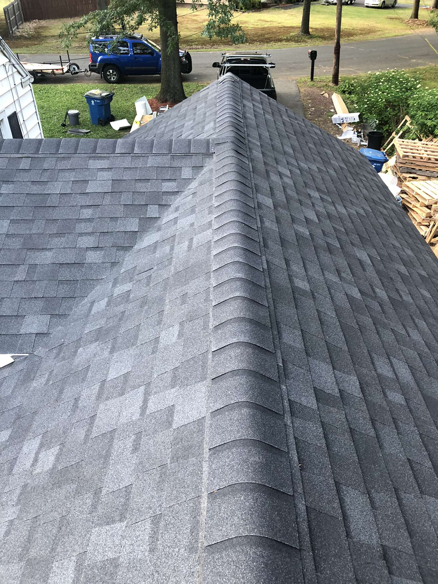 A high-angle view of a newly installed dark grey asphalt shingle roof on a house, with a residential street below.
