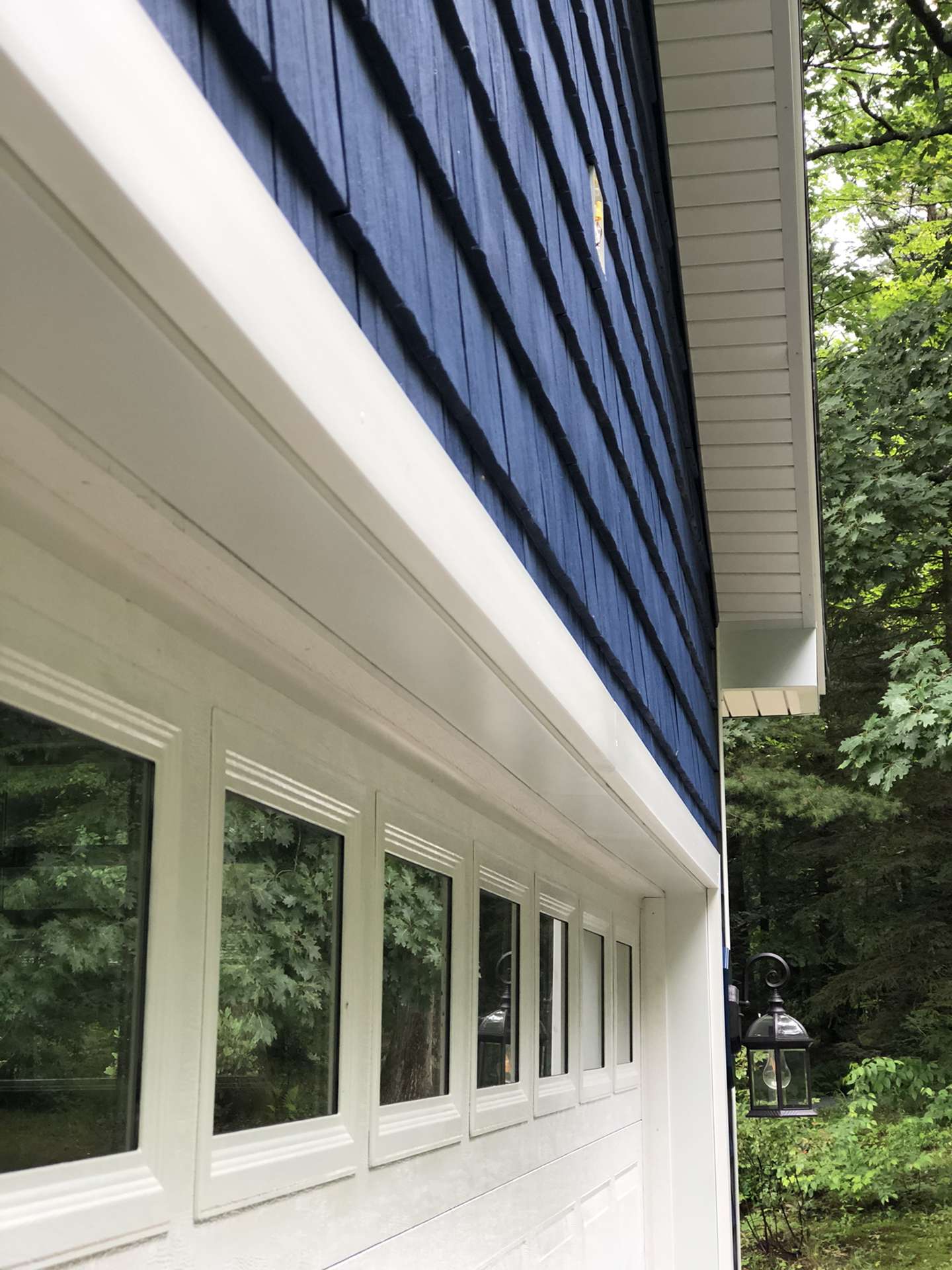 A low-angle view of a white garage door with windows under blue siding and a white soffit next to a forest.