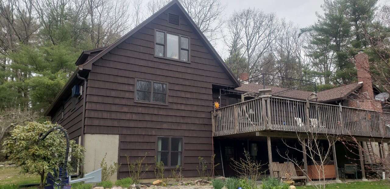 A multi-story, dark brown cedar shake house surrounded by trees with a wooden deck and a stone chimney.