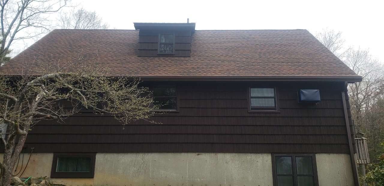 A two-story house with dark horizontal siding, a light-colored foundation, and a brown shingled roof with a dormer.