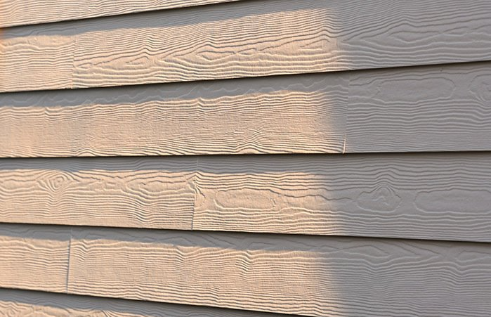 A close-up view of tan, wood-textured lap siding on an exterior wall, showing overlapping boards and a light shadow.