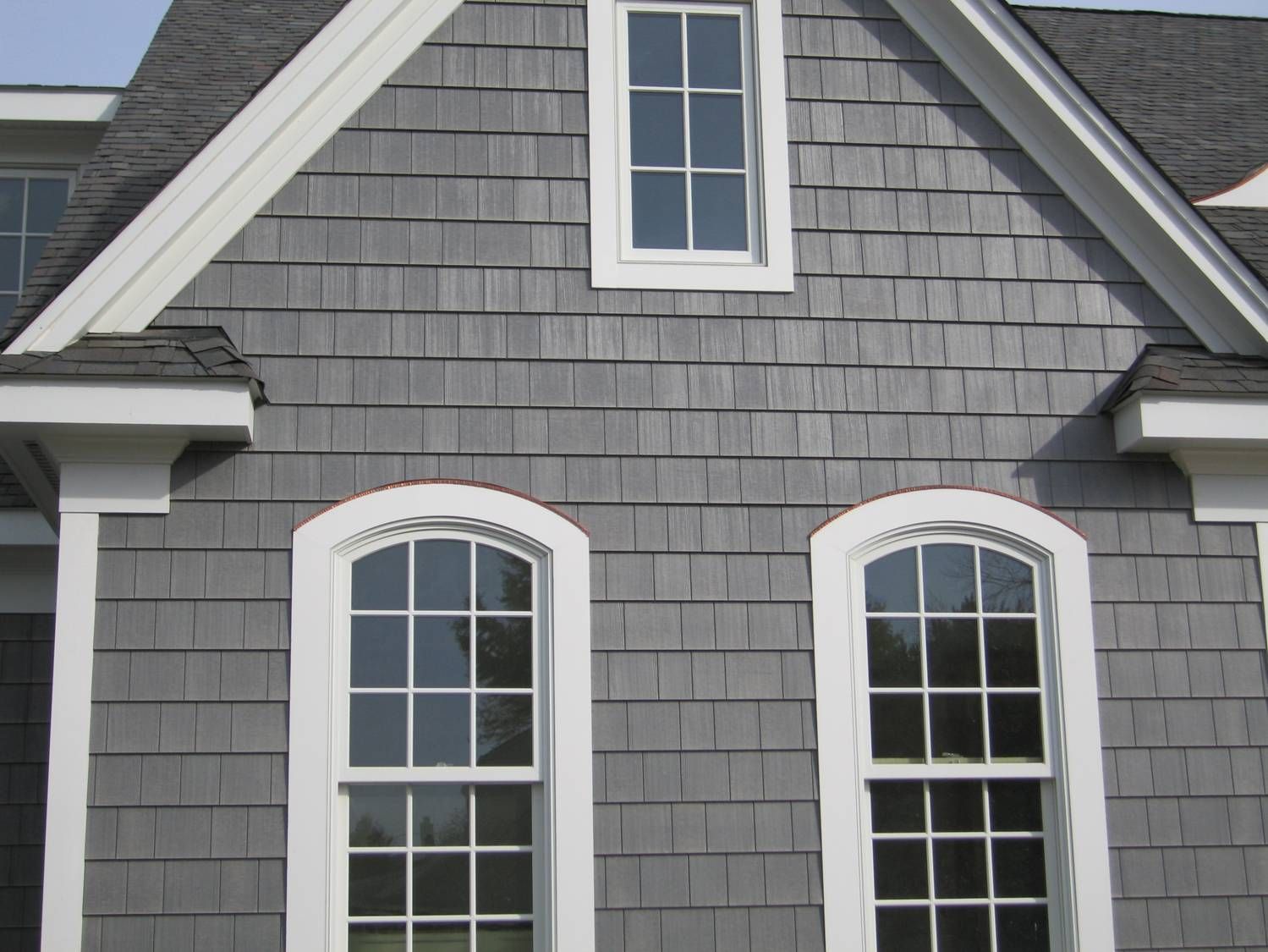 A gray shingled house exterior featuring a top centered rectangular window above two arched windows with white trim.