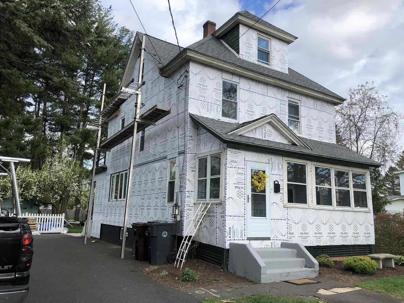 A multi-story house under renovation, covered in white insulating house wrap, with scaffolding against one side.