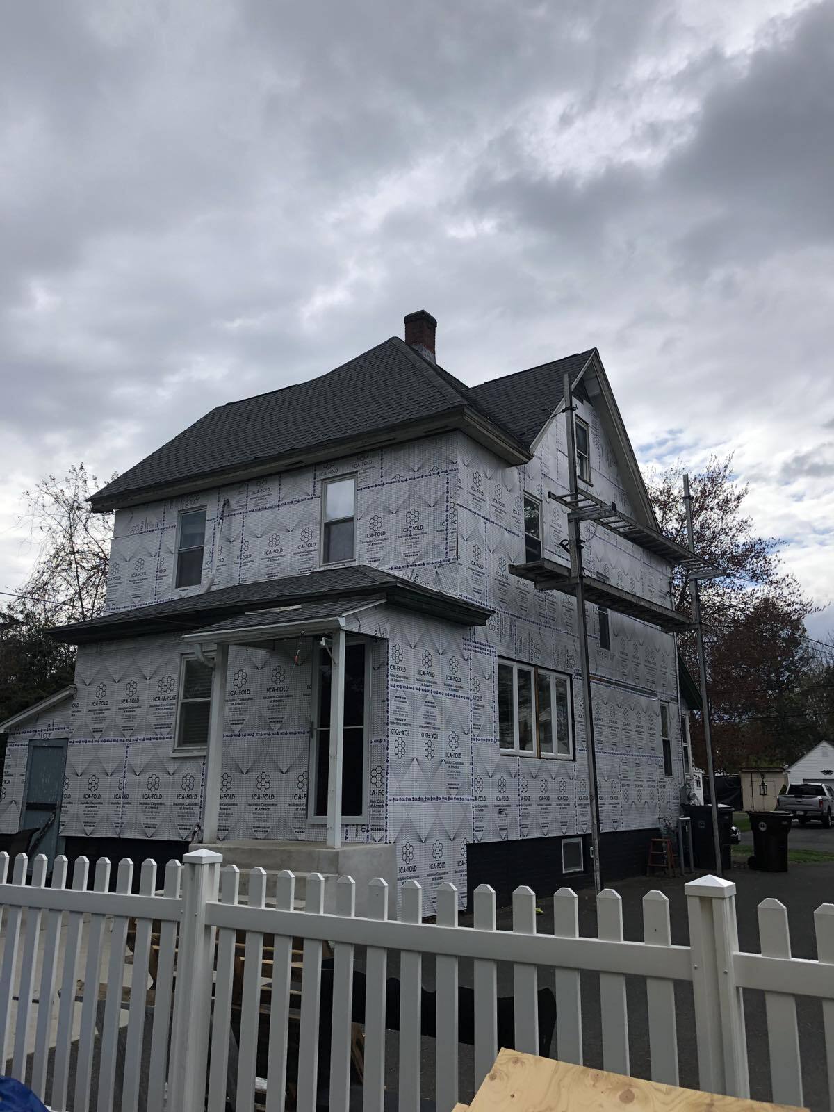 A two-story house under construction, covered in white house wrap, viewed behind a white picket fence on a cloudy day.