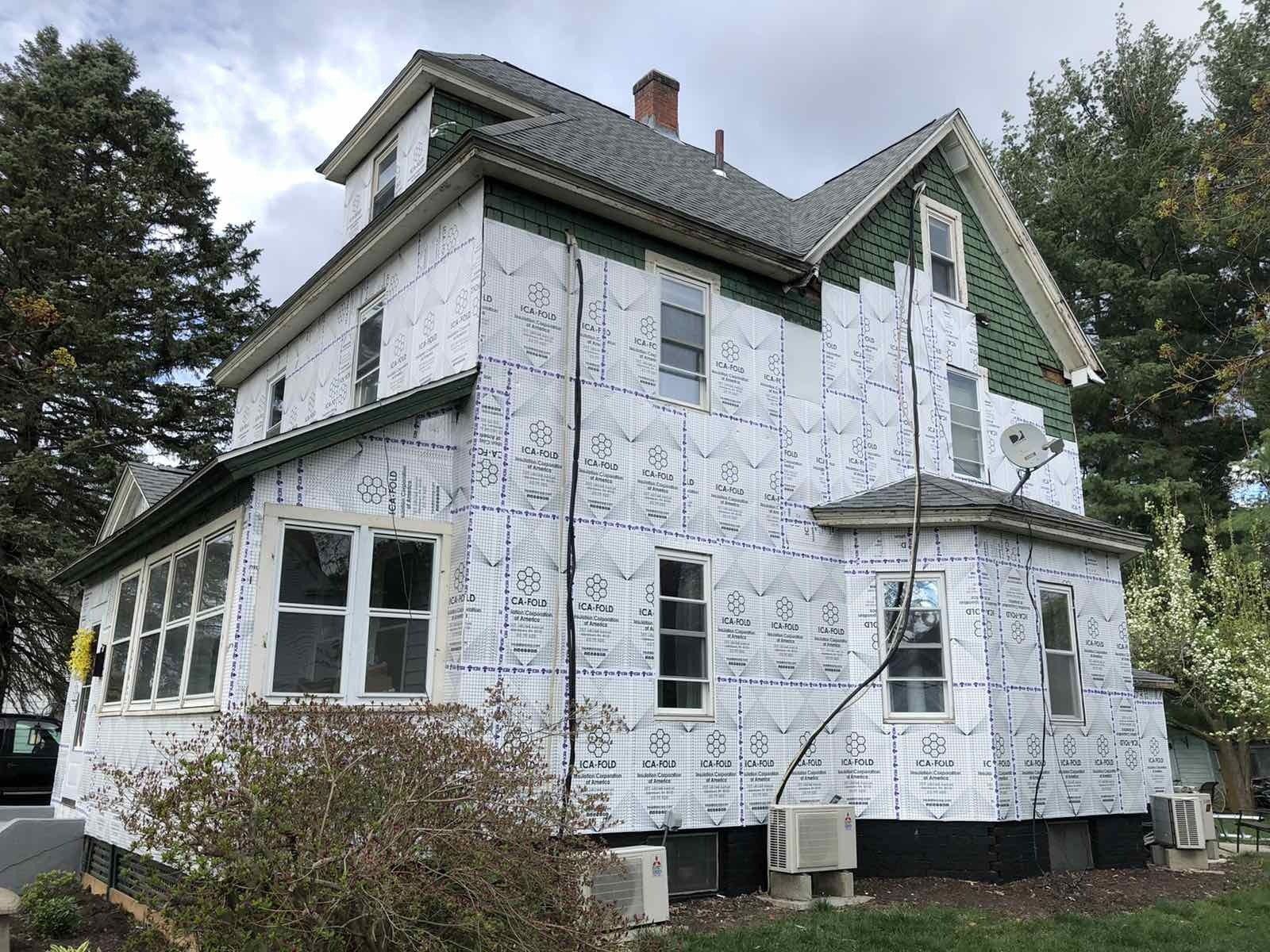 A two-story house under renovation, its exterior covered in white, grid-patterned insulation wrap and dark green trim.