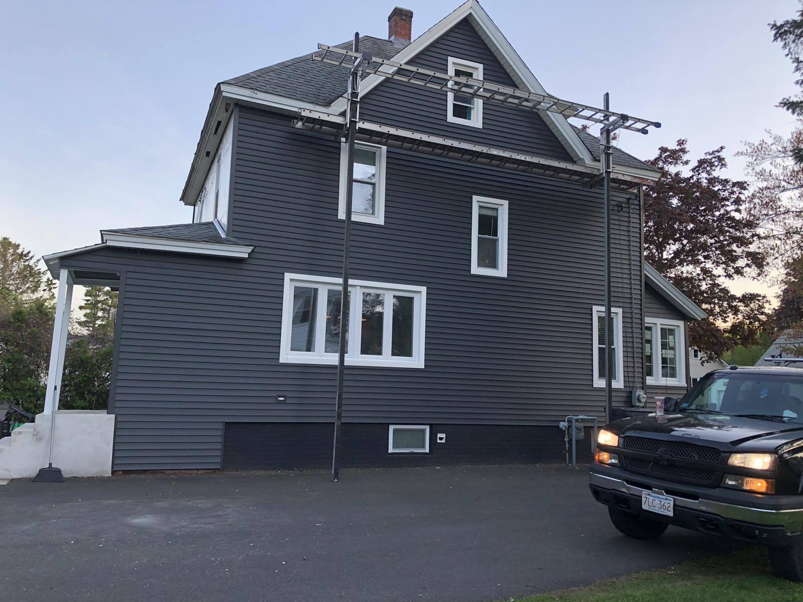A dark gray, two-story house with white trim stands beside a paved driveway with a black truck parked in front.