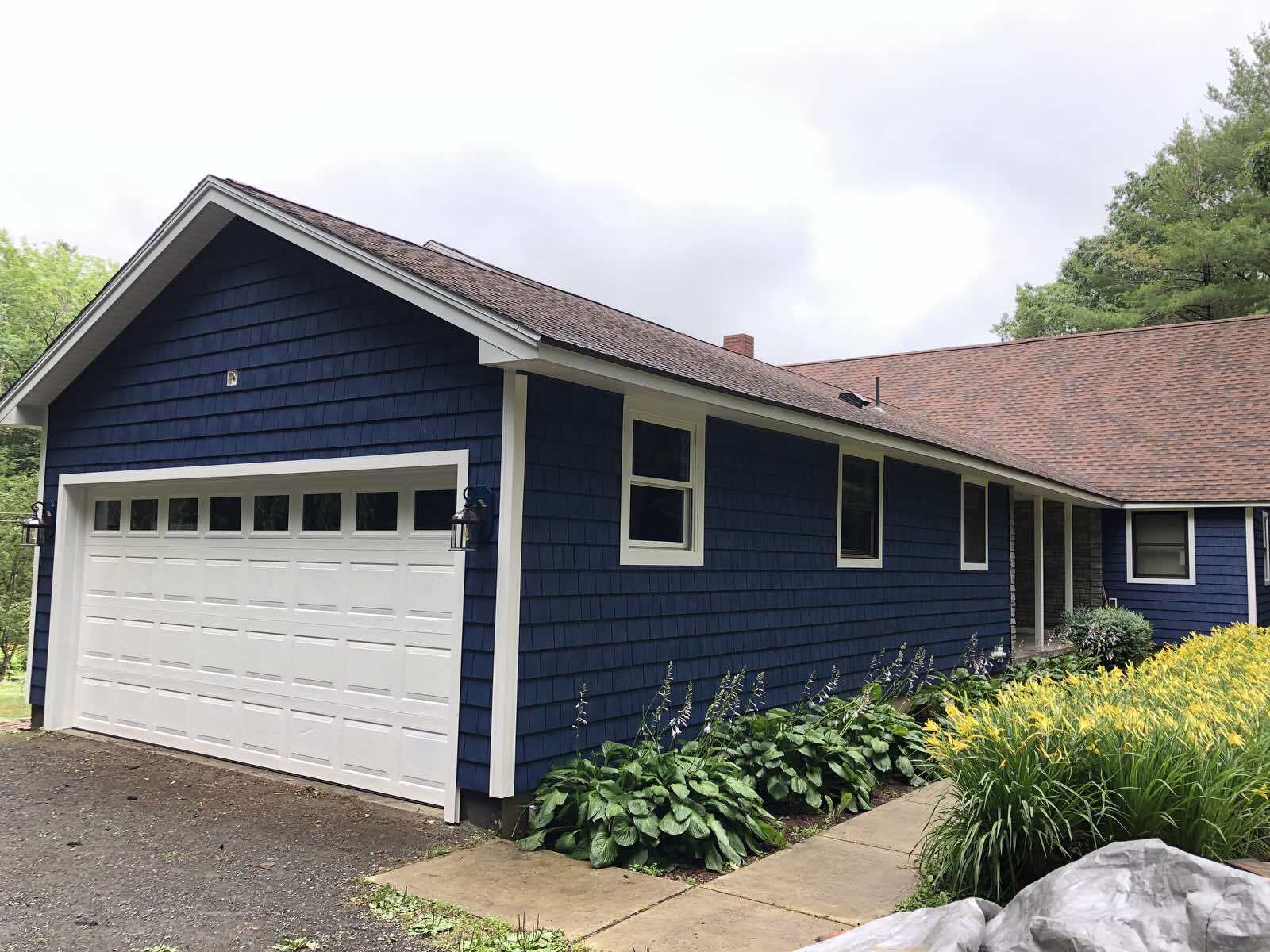 A dark blue house with white trim, a white garage door, and a brown shingled roof, set next to a walkway and garden.