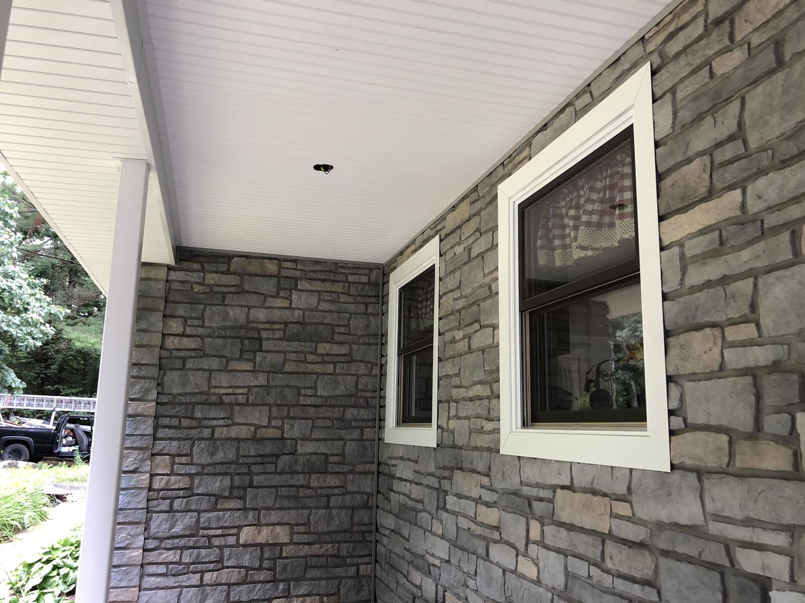 Exterior stone-veneer wall featuring two windows with white frames under a covered porch with a white ceiling.
