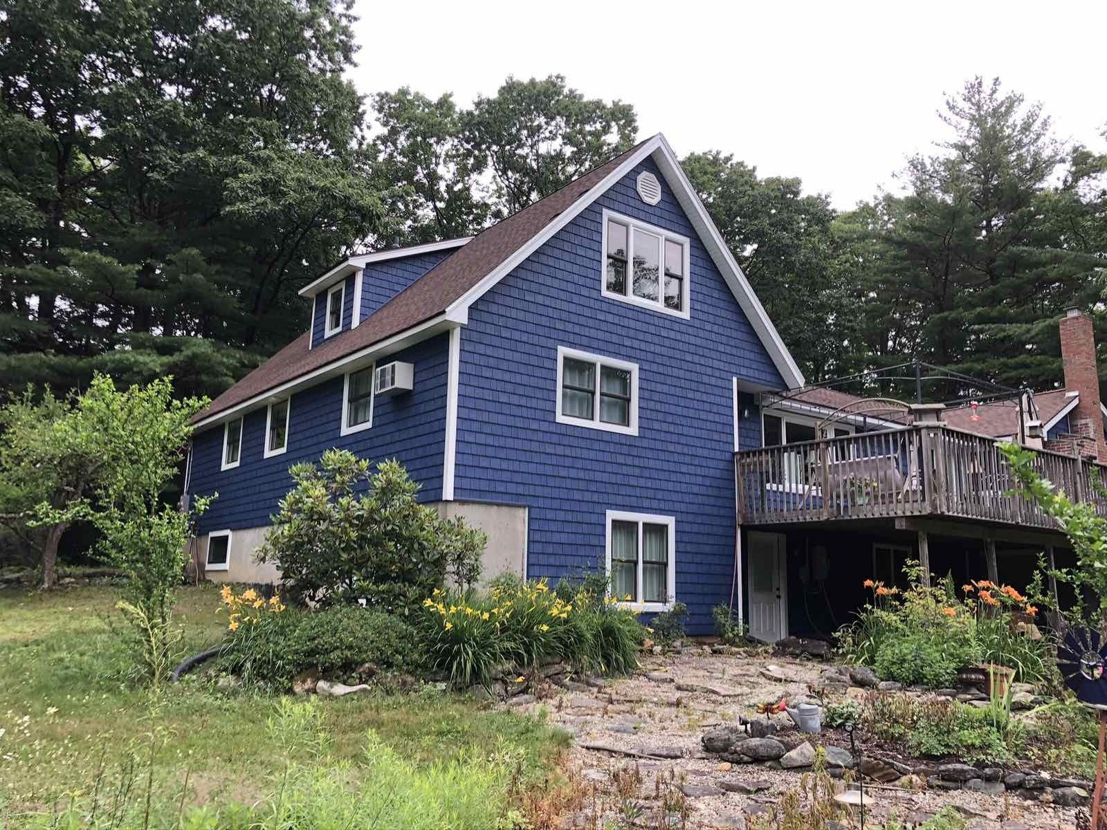 A two-story blue house with white trim and a wooden deck, situated among trees with a rocky garden in the foreground.