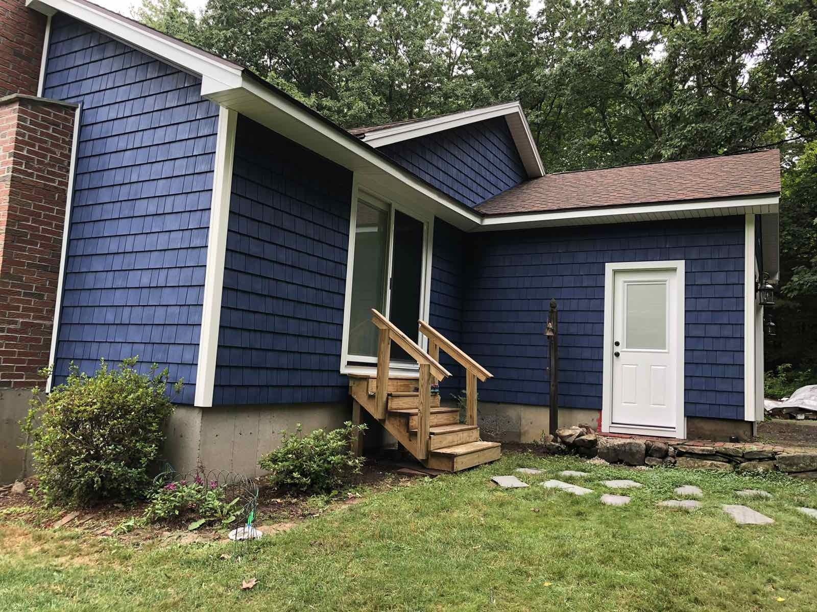 A blue house with cedar shake siding, white trim, a brick chimney, wooden stairs to a glass door, and a white side door.