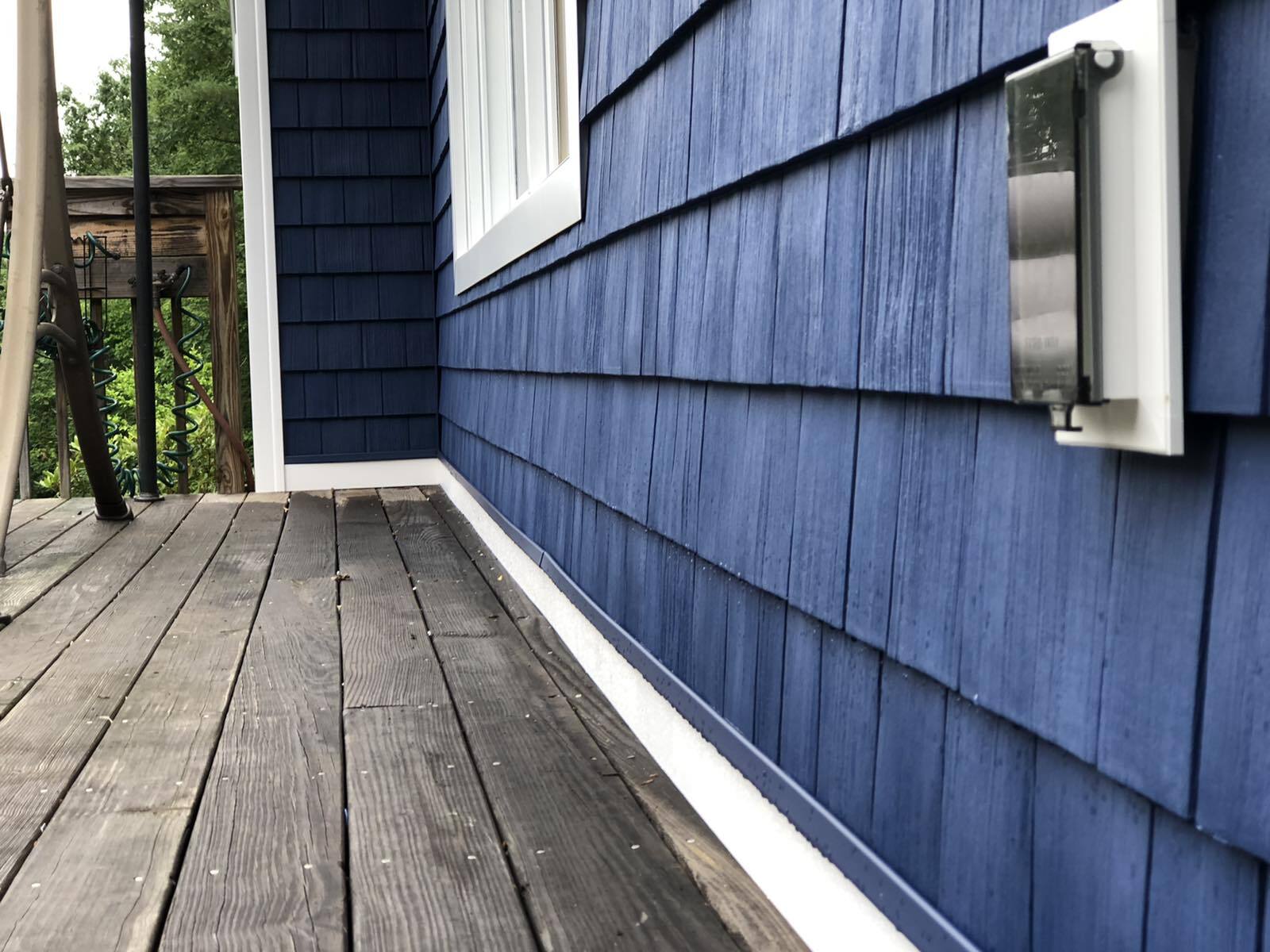 A wooden deck next to a house exterior featuring blue shingle siding, a white trim base, and a vertical wall-mounted box.