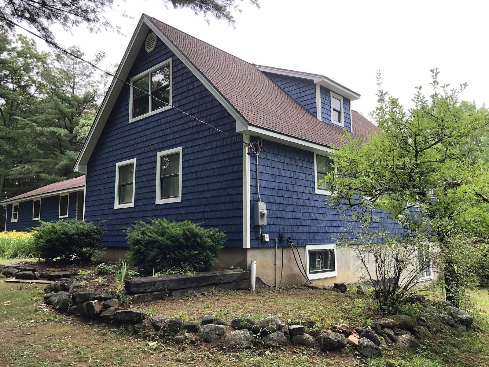 A two-story house with blue shake siding and a gabled roof, nestled in a grassy yard with a small stone retaining wall.