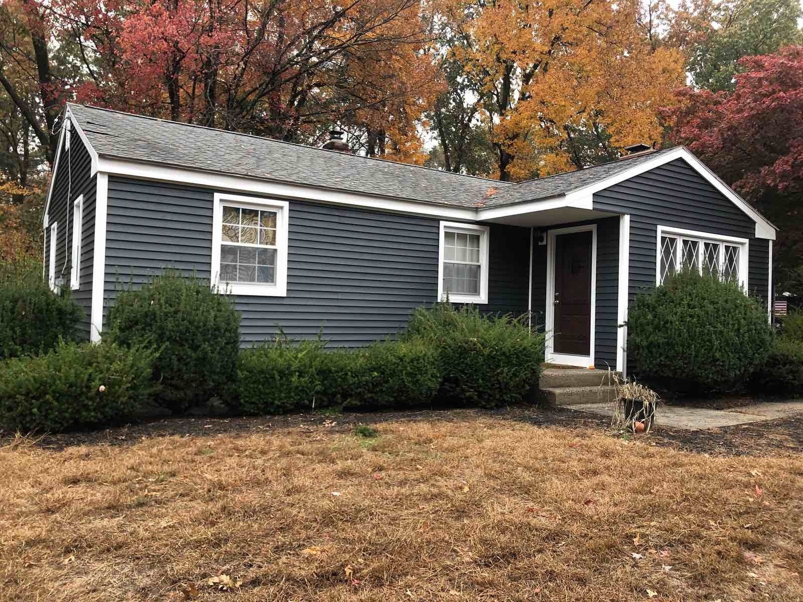 A single-story house with dark grey siding and white trim, surrounded by bushes and trees with autumn foliage.