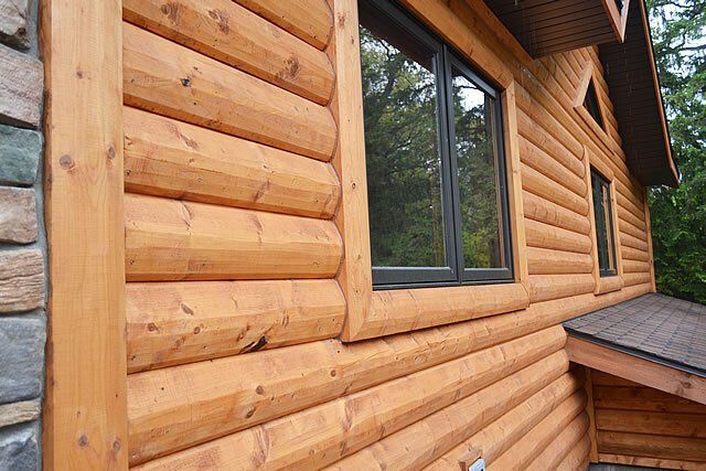 Close-up view of log cabin siding made of light-colored wood, featuring dark-framed windows and stone masonry.