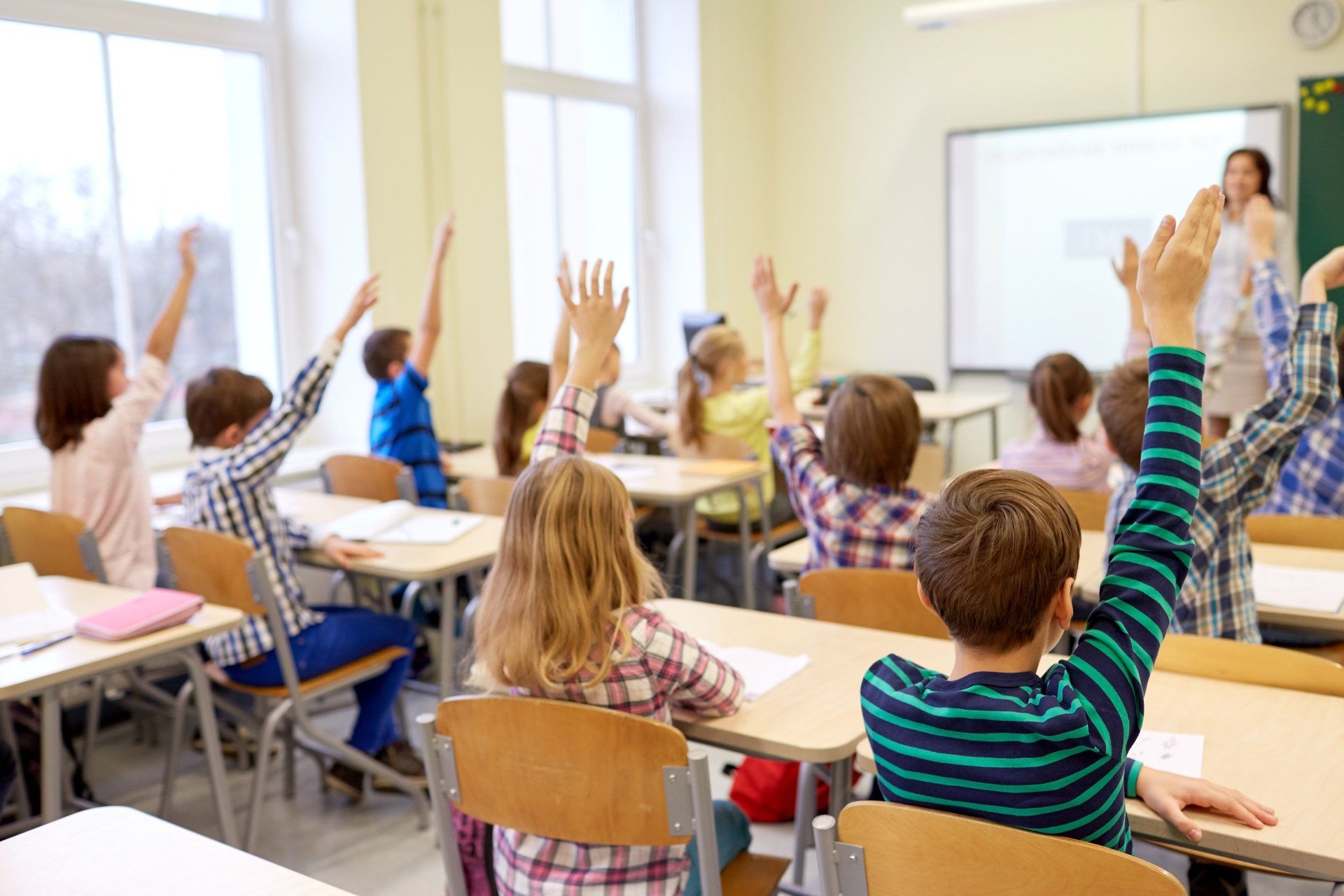 Students raising hands in a classroom, teacher at the front.