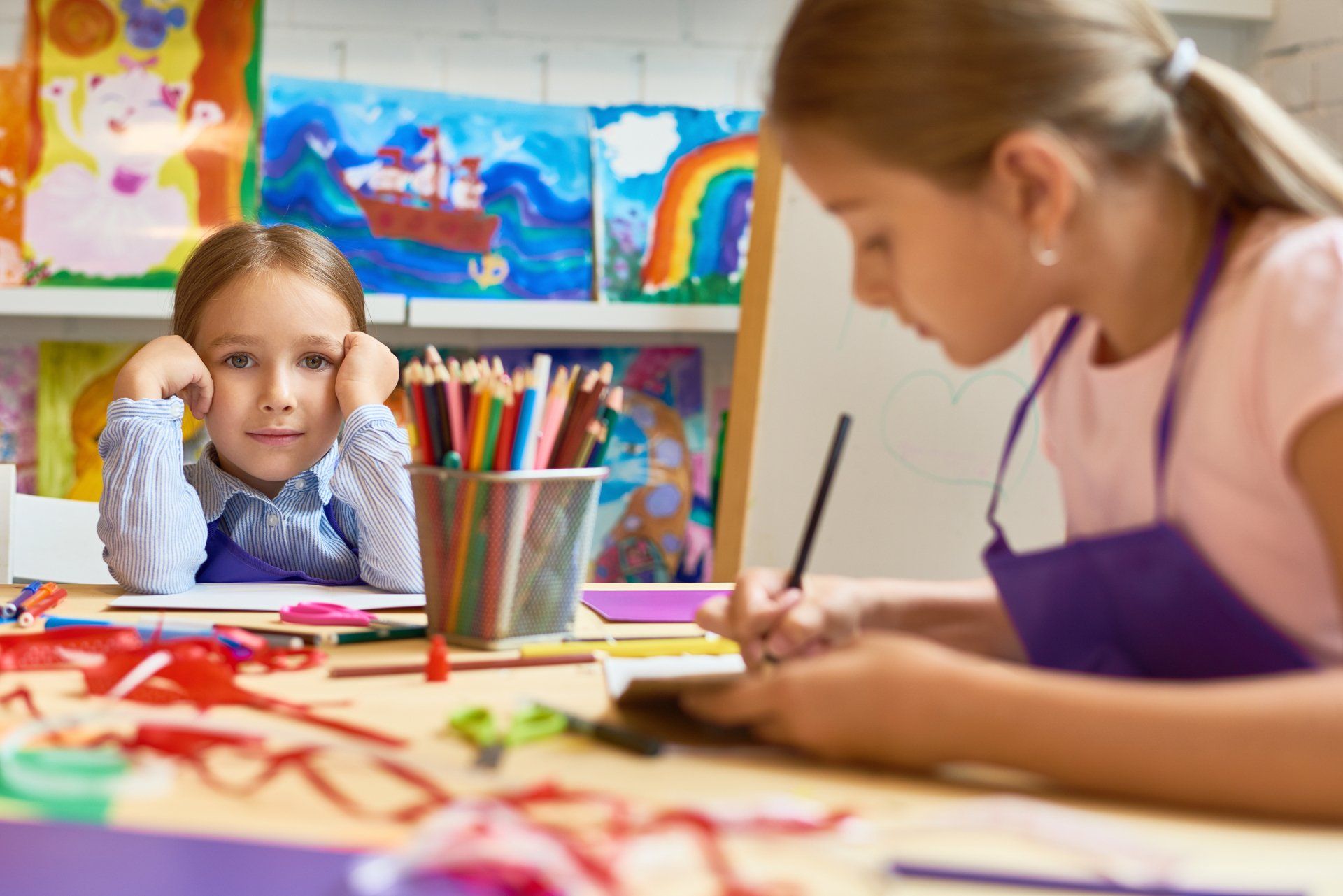 Two young girls in an art class, one drawing, the other looking at the viewer with a smile, surrounded by art supplies.
