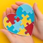 Hands holding a heart-shaped puzzle, in red, yellow and blue, on a yellow background.
