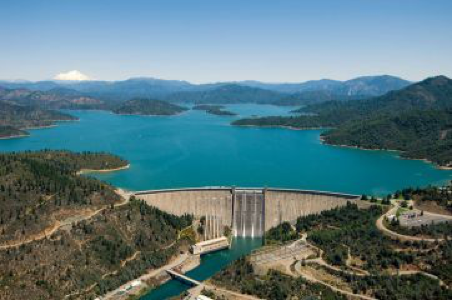 Aerial view of Shasta Dam with open spillways releasing water into river, surrounded by lush forest, mountainous landscape.