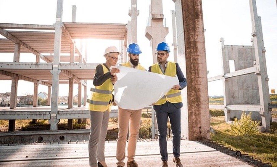Three construction pros in hard hats and vests discuss blueprints at site.