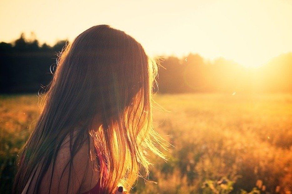 Young woman gazes across sunlit field at dusk, hair illuminated by setting sun.