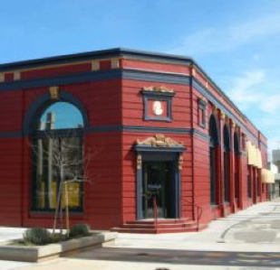 Red and black brick corner building with large windows, entrance canopy, and street-side trees under a clear blue sky.