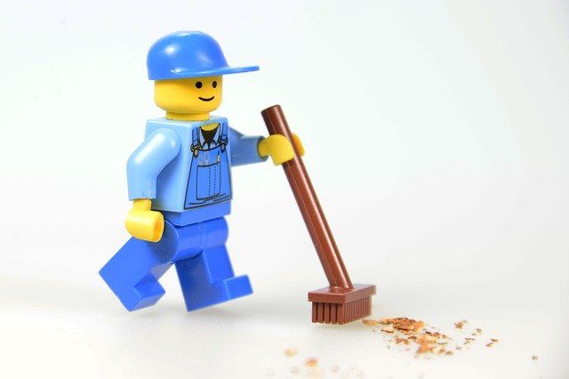 Lego figure in blue cap and overalls sweeping crumbs off floor with brown broom, against white background.
