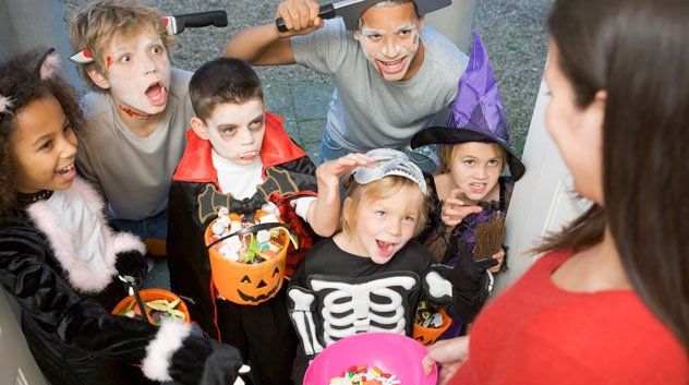 Children in Halloween costumes, including vampire and skeleton, collecting candy from a woman at a doorstep.