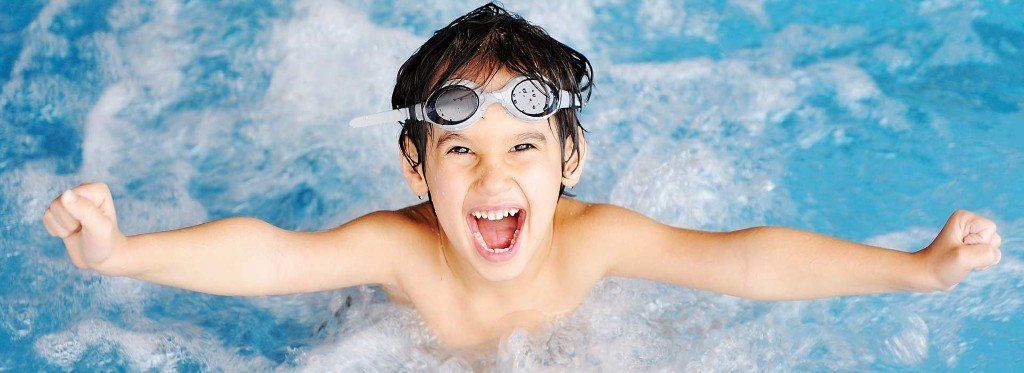 Boy swimming in a blue pool, arms outstretched, wearing goggles, mouth open in an excited expression.