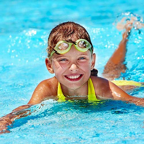 Girl swimming in blue pool, wearing goggles and yellow swimsuit, smiling.