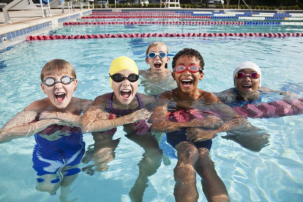Five kids in a swimming pool, wearing goggles and swim caps, smiling and laughing.