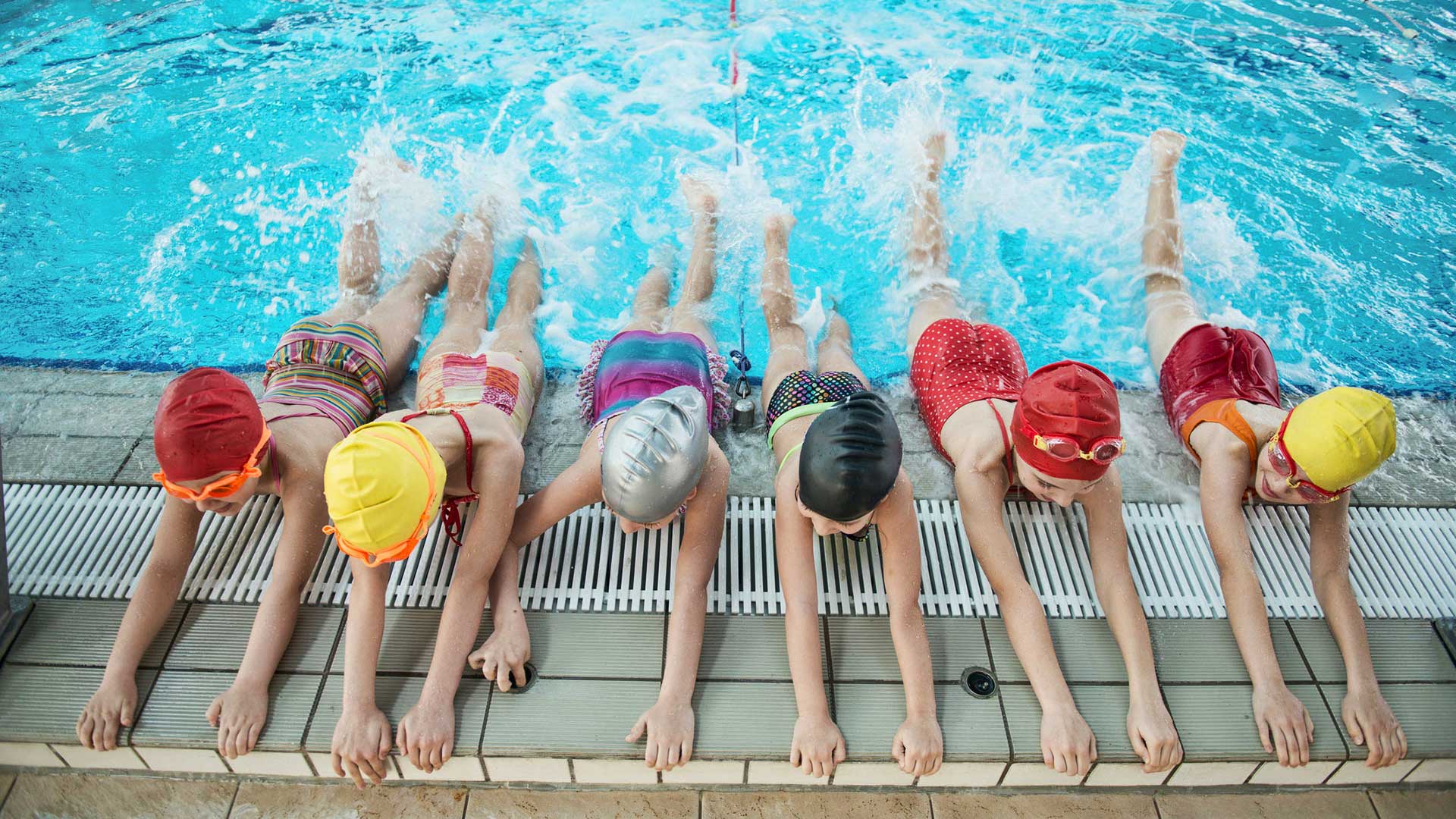 Swimmers in colorful caps and swimsuits, lined up at a pool edge, kicking in the water.