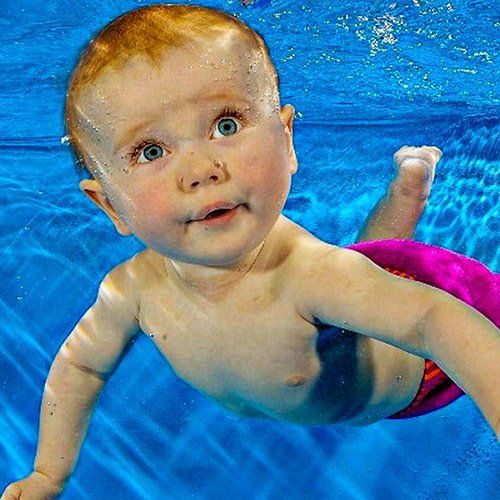 Baby swimming underwater, looking up with wide eyes; blue water, pink swim diaper.