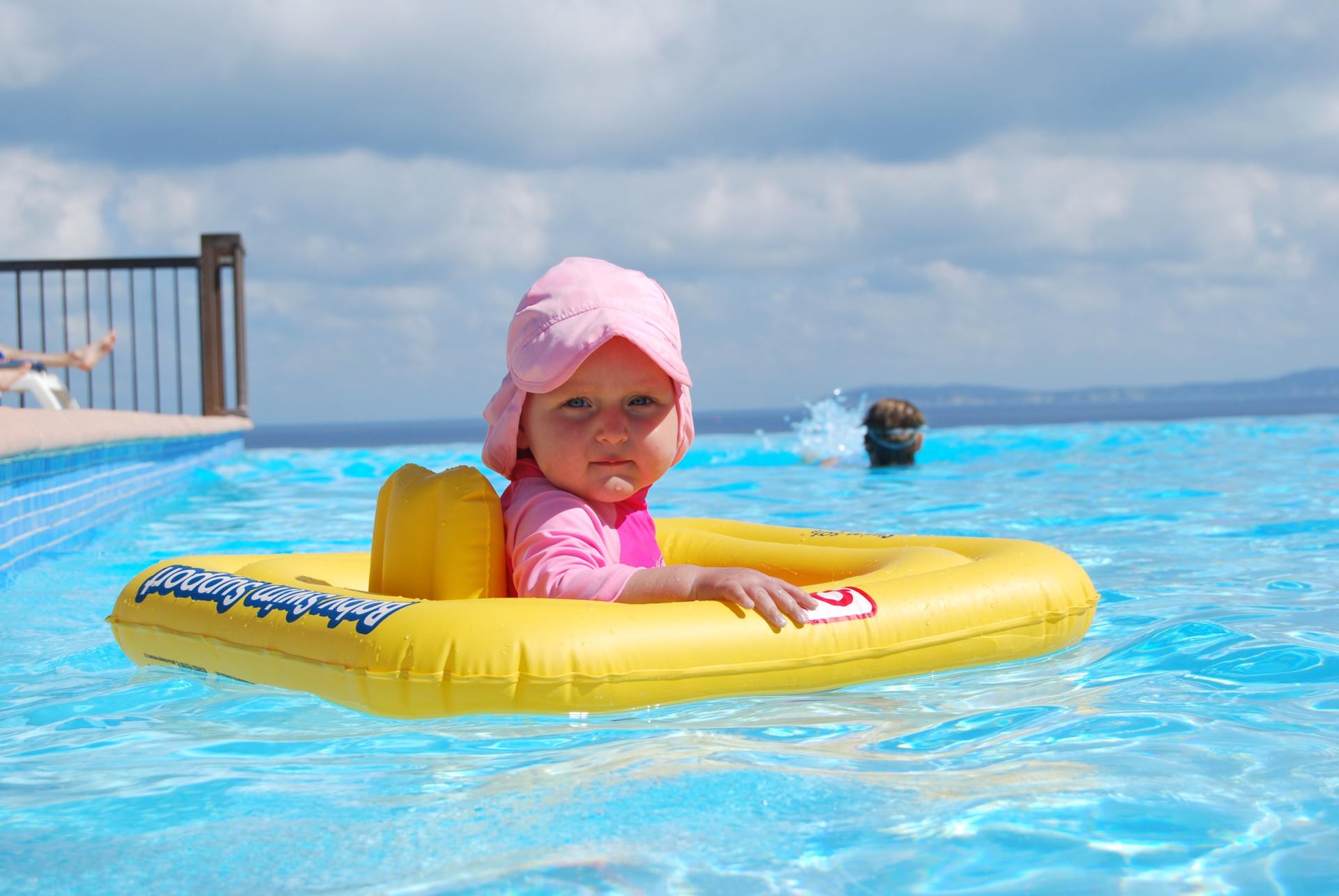 Child in a yellow inflatable boat in a swimming pool, wearing a pink hat and swim top, looking at the camera.
