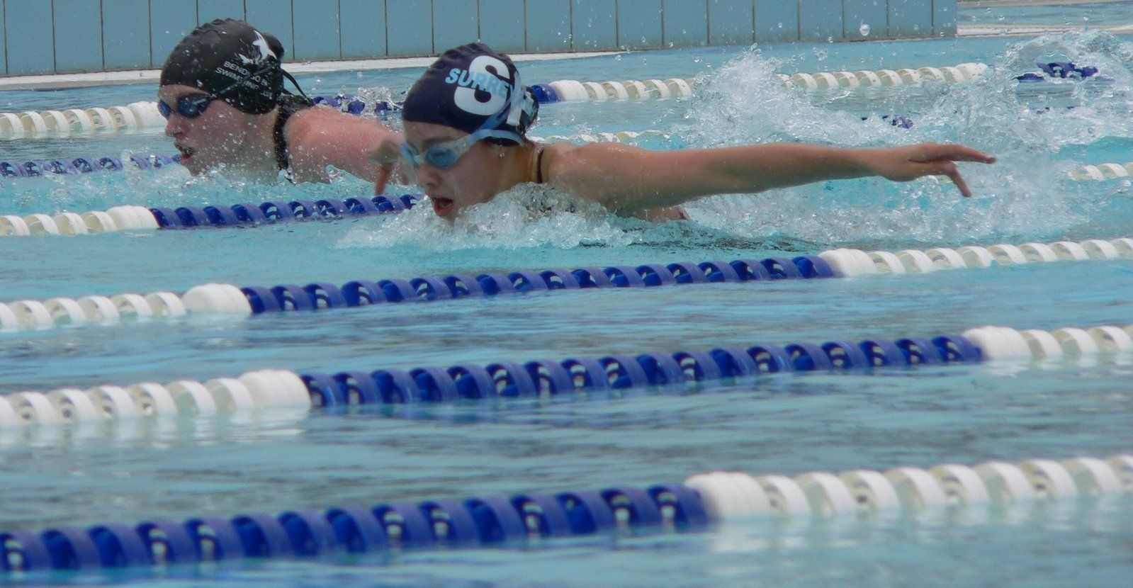 Two swimmers in a pool, butterfly stroke. One has dark hair, the other light hair.