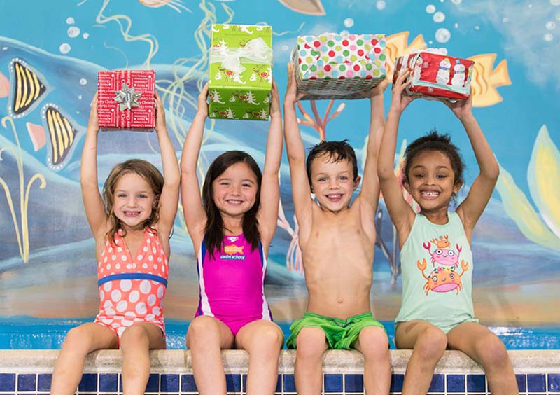 Four children in swimsuits hold up gift boxes near a pool, smiling.