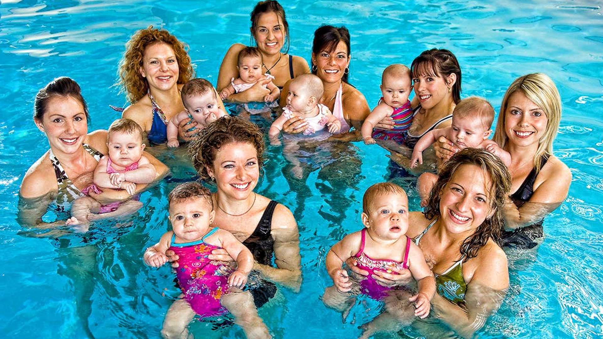 Mothers and babies in a swimming pool, smiling. Several babies are held in the water.