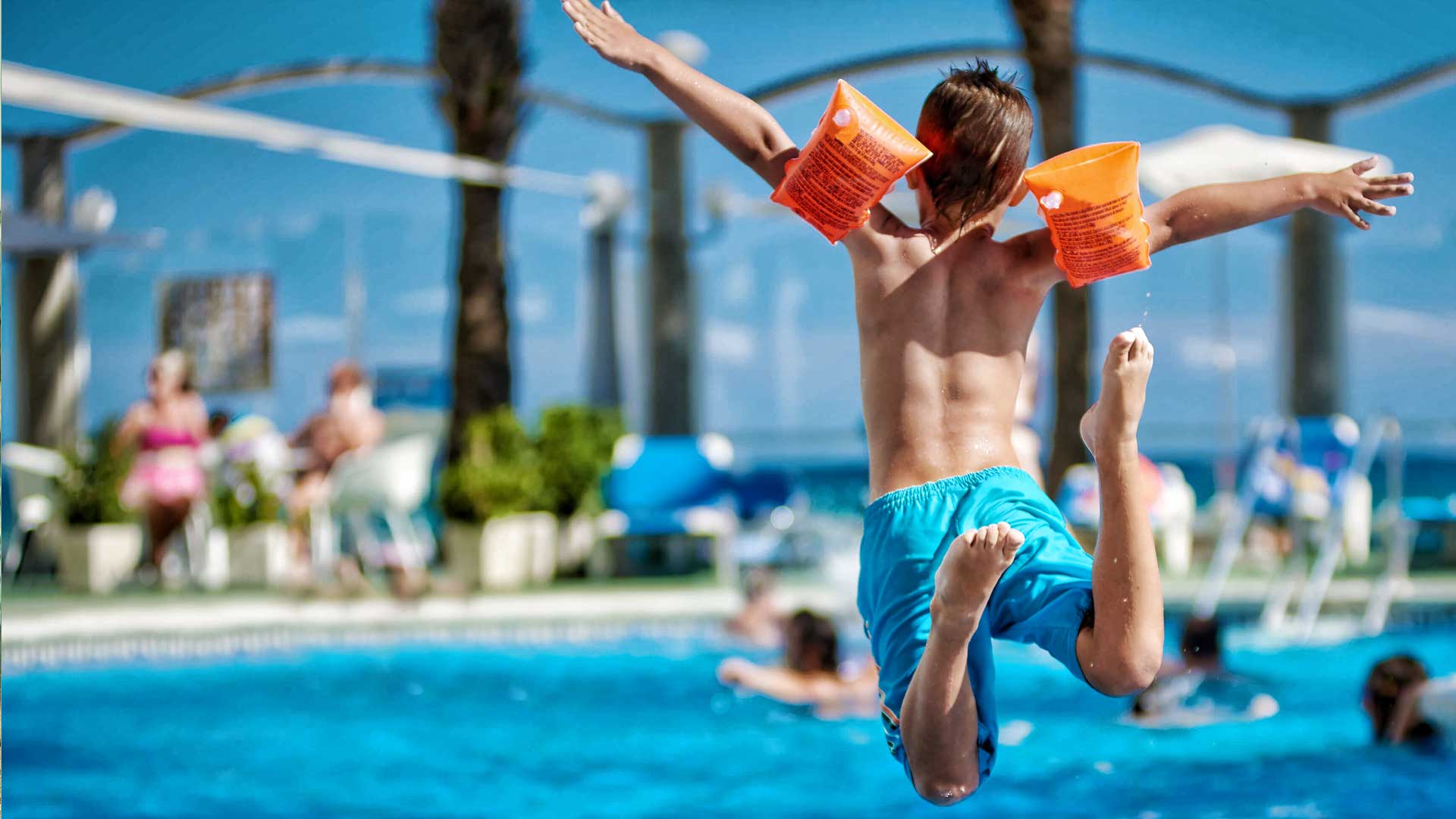 Boy jumping into a pool with orange arm floaties, arms outstretched, blue shorts, sunny outdoor setting.