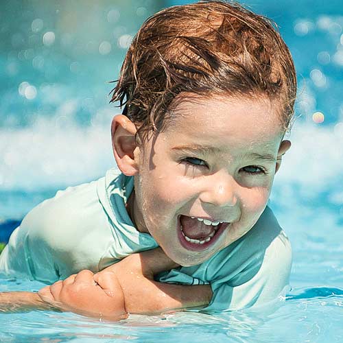 Young child in a swimming pool, smiling with mouth open, wearing a light blue swim shirt.