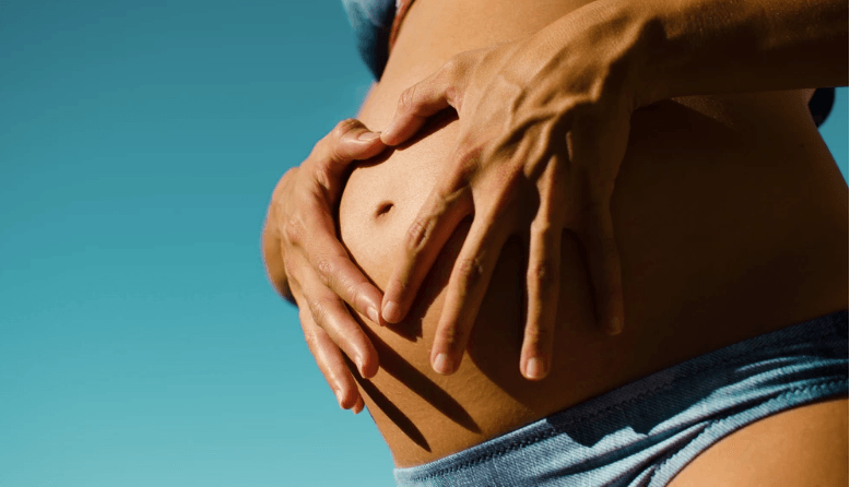 Pregnant person's belly, hands forming a heart shape. Blue sky background.