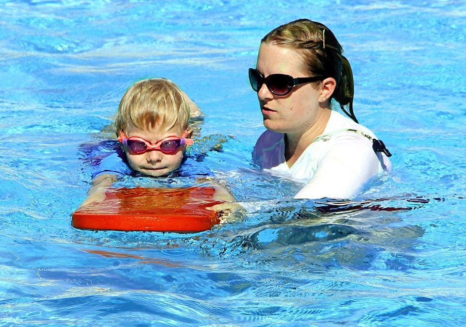 Child learning to swim with instructor, using a kickboard in a pool.