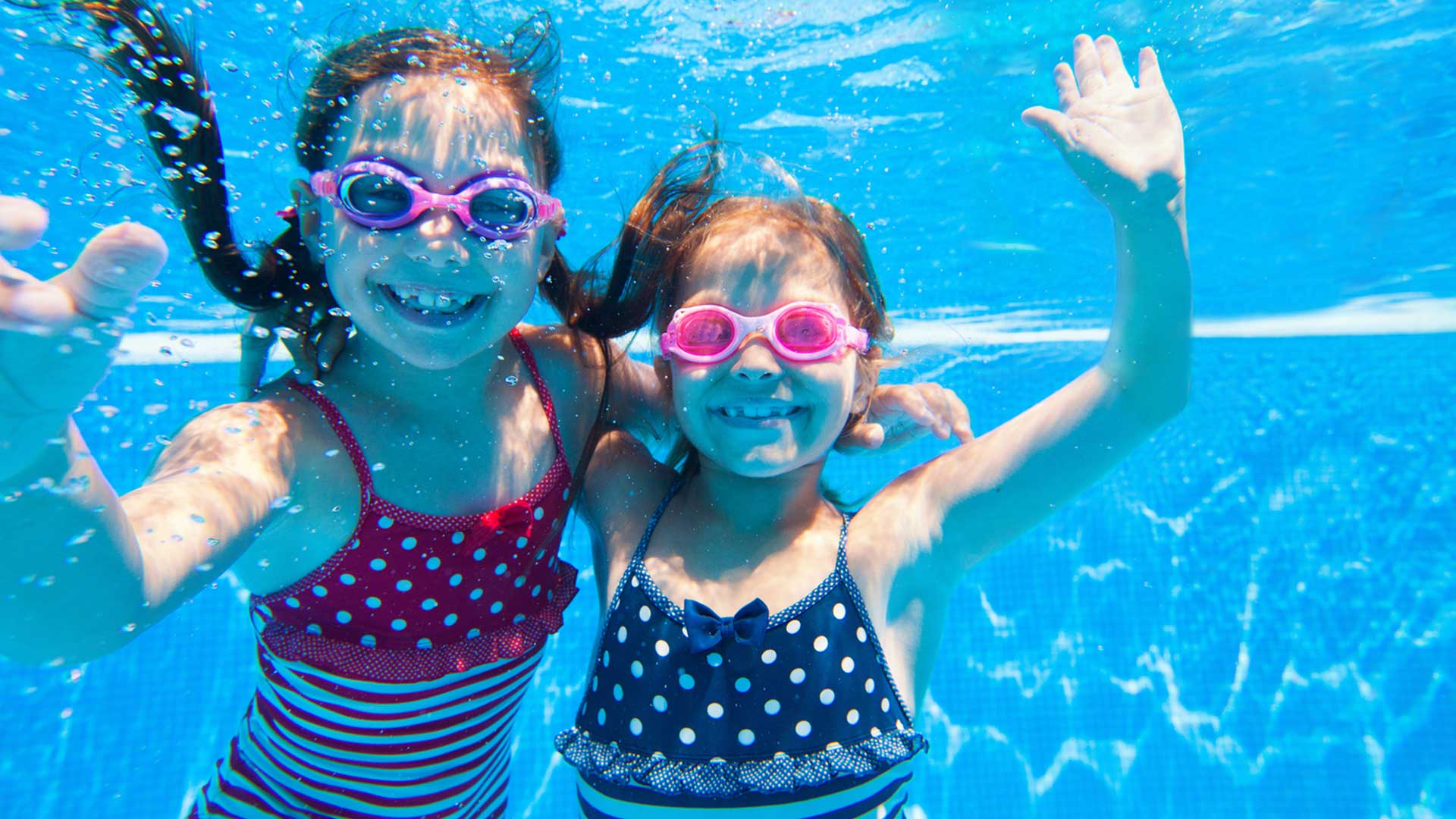 Two children underwater, smiling and wearing swim goggles, in a blue pool. One waves.