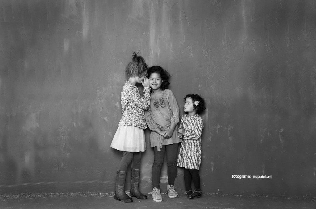 Drie kinderen staan ​​dicht bij elkaar voor een effen muur, twee van hen dragen jurken met een patroon.